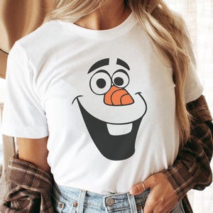 May include: White t-shirt featuring a cartoon face of Olaf from Frozen. The design includes black eyes, a carrot nose, and a large, smiling mouth. The shirt is a crew neck style.