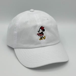 May include: White baseball cap featuring an embroidered Minnie Mouse design. The Minnie Mouse graphic includes a red bow, black head and body, and a red and white polka dot dress. The cap has a curved brim and a top button.