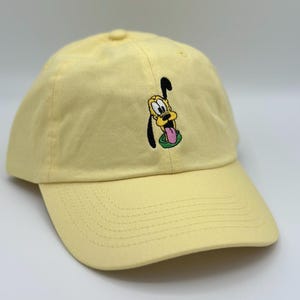 May include: A light yellow baseball cap featuring an embroidered image of Pluto, the Disney cartoon dog. The cap has a curved brim and an adjustable strap. The Pluto image includes black, yellow, pink, and green colors.