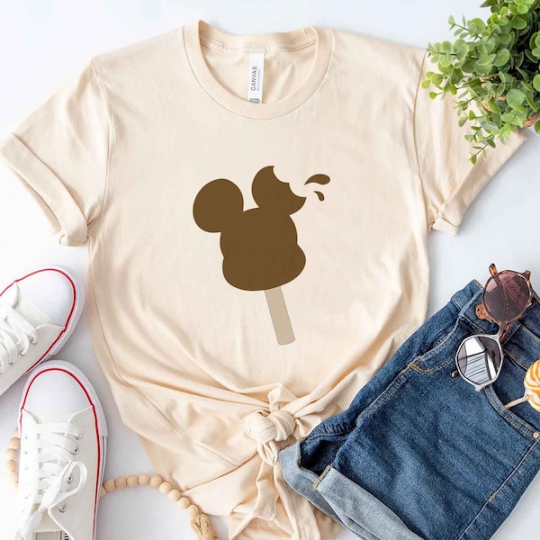 Könnte beinhalten: Ein beiges T-Shirt mit einem braunen Mickey Mouse Eiscreme-Motiv. Das Eis ist auf einem Stiel und hat einen Bissen.