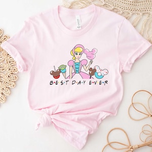 May include: Pink t-shirt with a cartoon image of a princess, Mickey Mouse ears, a green alien, a Mickey Mouse ice cream cone, and a Mickey Mouse donut. The text "BEST DAY EVER" is printed below the image.