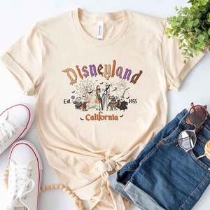 May include: A beige t-shirt with a graphic print of the Disneyland logo and characters from the Nightmare Before Christmas. The text reads "Disneyland Est. 1955 California".