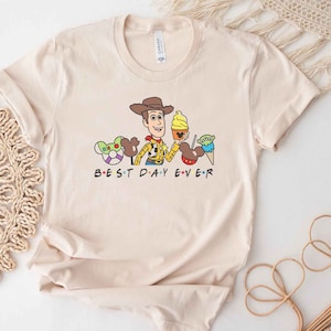 May include: A beige t-shirt with a cartoon image of Woody from Toy Story, Mickey Mouse ears, and a green alien. The text "BEST DAY EVER" is printed below the image.