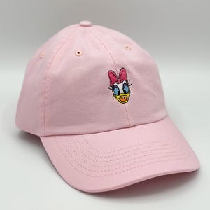 May include: A light pink baseball cap featuring an embroidered Daisy Duck design on the front. The cap has a curved brim and an adjustable strap. The Daisy Duck embroidery includes a pink bow, blue eyes, and a yellow beak.