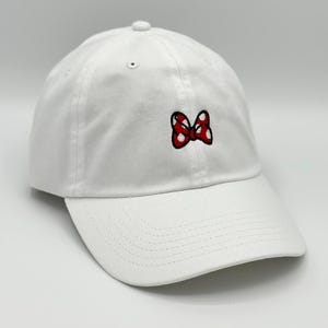 May include: White baseball cap with a red and white polka dot bow embroidered on the front. The cap has a curved brim and a classic six-panel design. A casual accessory for everyday wear.