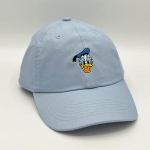 May include: A light blue baseball cap featuring an embroidered image of Donald Duck. The cap has a curved brim and an adjustable strap. The Donald Duck image is in yellow, blue, and white.