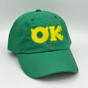 May include: Green baseball cap with the word "OK" embroidered in yellow. The hat has a curved brim and a rounded crown. The text is in a bold, playful font.