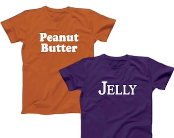 May include: Two short-sleeved t-shirts. One is orange with the text "Peanut Butter" in white. The other is purple with the text "Jelly" in white. Both shirts are made of a soft material.