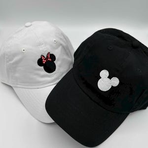 May include: Two baseball caps, one white and one black. The white cap features an embroidered Minnie Mouse design with a black head and a red and white polka dot bow. The black cap has a white embroidered Mickey Mouse design.