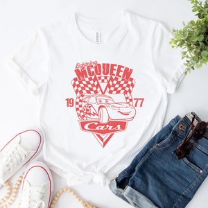 以下が含まれることがあります： 映画「カーズ」のライトニング・マックイーンの赤いグラフィックがプリントされた白いTシャツ。グラフィックには、「ライトニング・マックイーン」、「19」、「77」、「カーズ」の文字が含まれています。