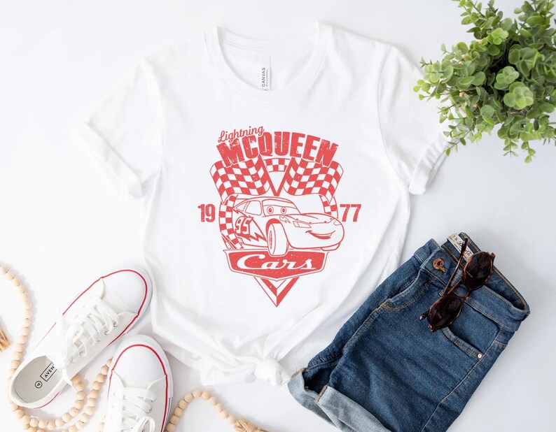以下が含まれることがあります： 映画「カーズ」のライトニング・マックイーンの赤いグラフィックがプリントされた白いTシャツ。グラフィックには、「ライトニング・マックイーン」、「19」、「77」、「カーズ」の文字が含まれています。