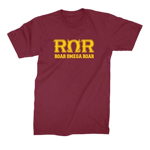 Peut inclure: Un t-shirt bordeaux avec un motif dor&eacute; qui dit "ROR" et "ROAR OMEGA ROAR".
