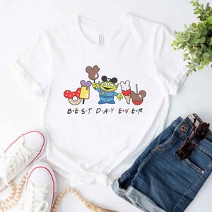 May include: White t-shirt with a cartoon image of a green alien character holding a Mickey Mouse ice cream cone and a donut. The text "BEST DAY EVER" is printed below the image.