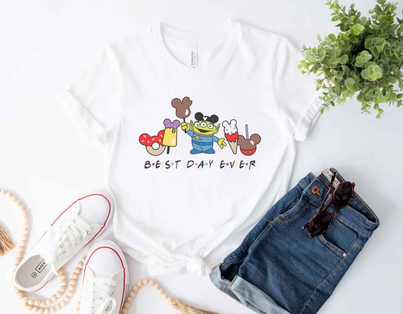 May include: White t-shirt with a cartoon image of a green alien character holding a Mickey Mouse ice cream cone and a donut. The text "BEST DAY EVER" is printed below the image.