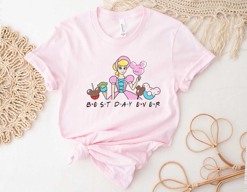 May include: Pink t-shirt with a cartoon image of a princess, Mickey Mouse ears, a green alien, a Mickey Mouse ice cream cone, and a Mickey Mouse donut. The text "BEST DAY EVER" is printed below the image.