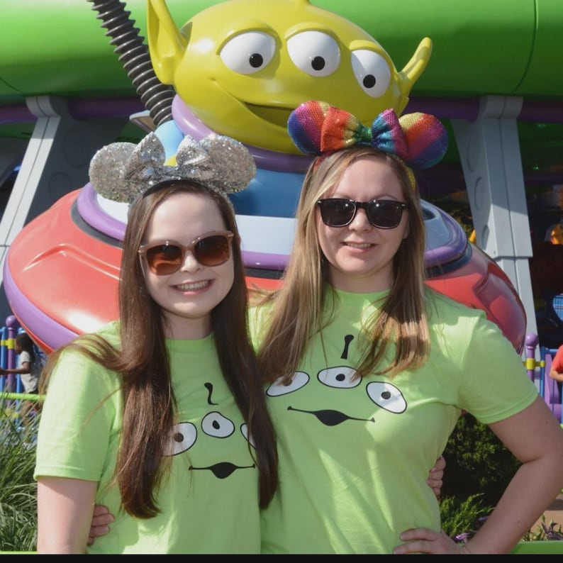 May include: Two people wearing lime green t-shirts with a three-eyed alien graphic, sunglasses, and mouse ear headbands. One headband is silver glitter, and the other is a rainbow bow. A large green alien statue is in the background.