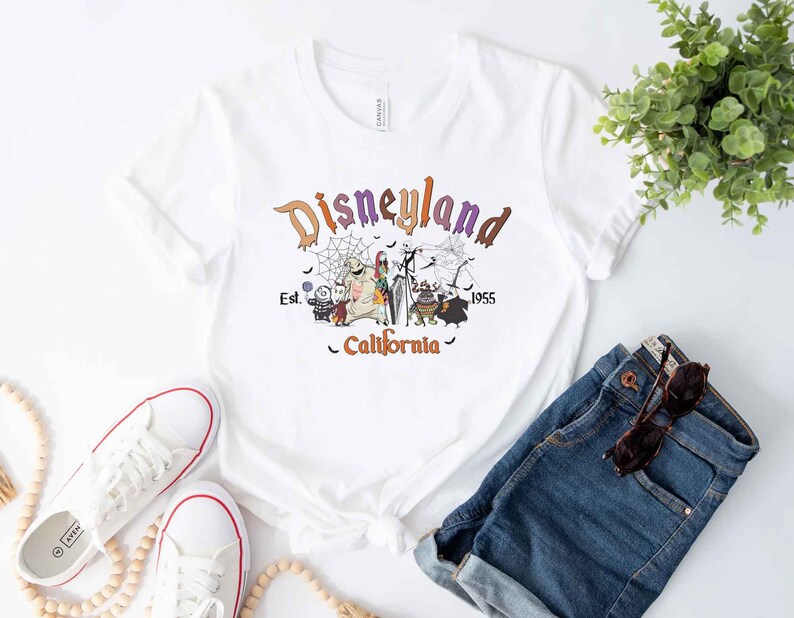 May include: White t-shirt with a purple, orange, and black graphic of the Nightmare Before Christmas characters in front of a Disneyland sign that reads "Disneyland Est. 1955 California".