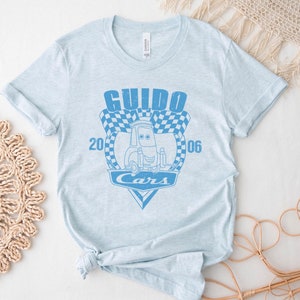 以下が含まれることがあります： 薄い青色のTシャツに、映画カーズのキャラクター、グイドの青いグラフィックがプリントされています。グラフィックには、「グイド」と「カーズ」の文字と、市松模様の旗のデザインが描かれています。