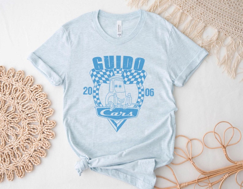 以下が含まれることがあります： 薄い青色のTシャツに、映画カーズのキャラクター、グイドの青いグラフィックがプリントされています。グラフィックには、「グイド」と「カーズ」の文字と、市松模様の旗のデザインが描かれています。