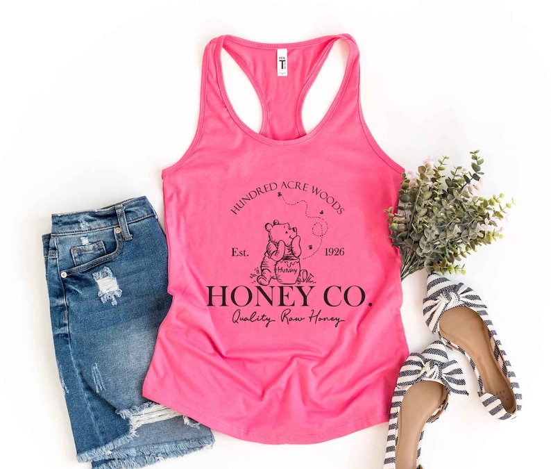 May include: Pink tank top with a black graphic of Winnie the Pooh and a honey pot. The graphic says "Hundred Acre Woods Est. 1926 Honey Co. Quality Raw Honey".