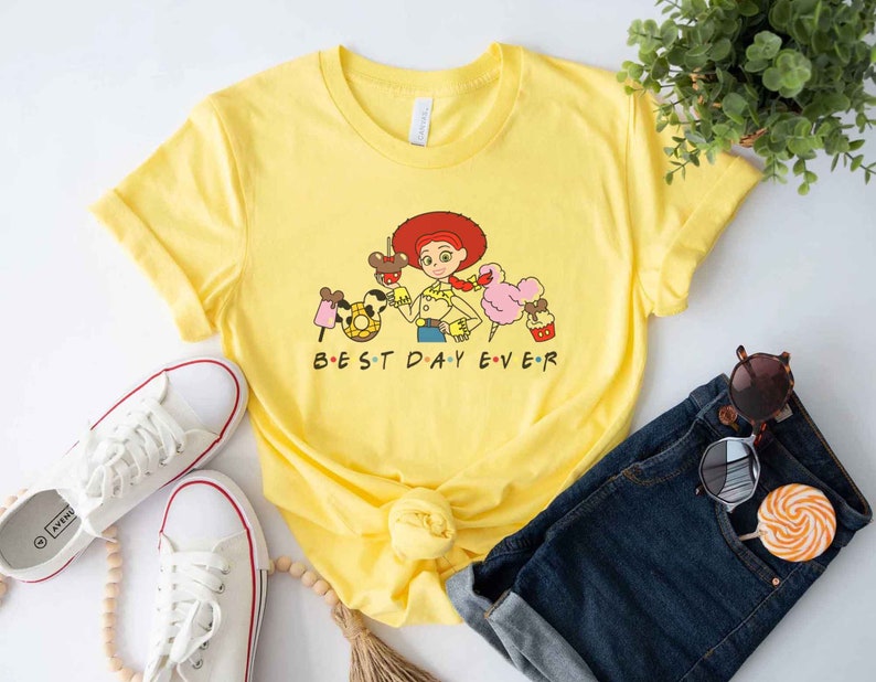 May include: Yellow t-shirt with a cartoon image of a character from the Toy Story movie franchise. The character is wearing a red and white cowboy hat and holding a pink cotton candy. The text "BEST DAY EVER" is printed on the shirt.