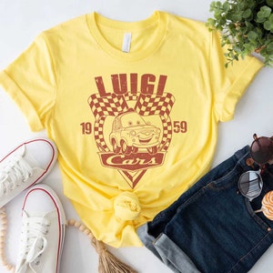 以下が含まれることがあります： 映画「カーズ」のルイージのヴィンテージスタイルのグラフィックがプリントされた黄色のTシャツ。グラフィックにはルイージの車、チェッカーフラッグ、そして「ルイージカーズ」というテキストと数字の19と59が描かれています。