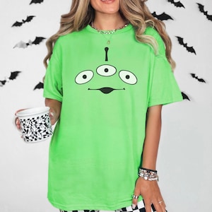 May include: A neon green t-shirt featuring a cartoon alien face with three eyes and a curved mouth. The shirt is paired with black and white checkered pants. A white mug with a black and white checkered pattern is held in the left hand.