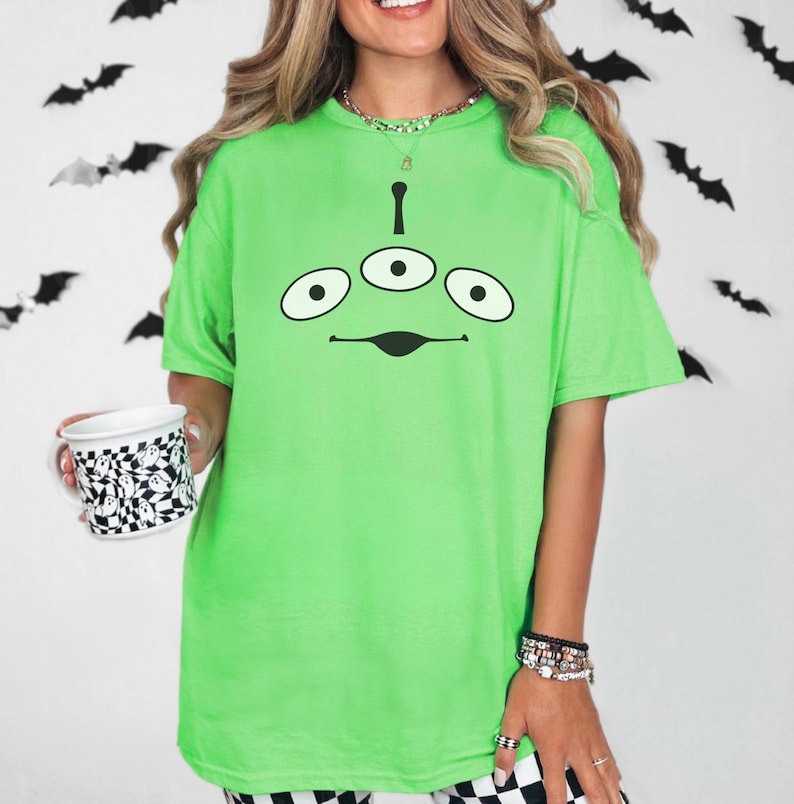May include: A neon green t-shirt featuring a cartoon alien face with three eyes and a curved mouth. The shirt is paired with black and white checkered pants. A white mug with a black and white checkered pattern is held in the left hand.