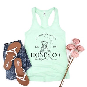 May include: A light green tank top with a black graphic of Winnie the Pooh holding a jar of honey. The graphic also includes the text "Hundred Acre Woods", "Est. 1926", "Honey Co.", and "Quality Raw Honey".