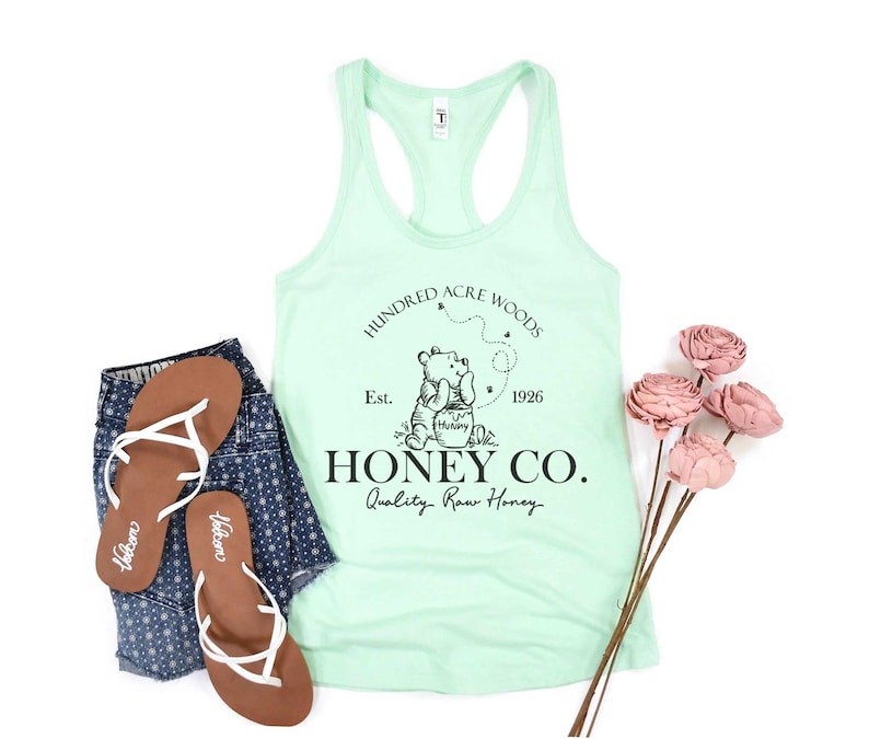 May include: A light green tank top with a black graphic of Winnie the Pooh holding a jar of honey. The graphic also includes the text "Hundred Acre Woods", "Est. 1926", "Honey Co.", and "Quality Raw Honey".