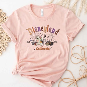 May include: A pink t-shirt with a vintage-style graphic of the Disneyland logo, featuring characters from the Nightmare Before Christmas. The text reads "Disneyland Est. 1955 California".