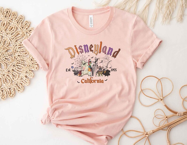 May include: A pink t-shirt with a vintage-style graphic of the Disneyland logo, featuring characters from the Nightmare Before Christmas. The text reads "Disneyland Est. 1955 California".