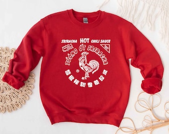 May include: Red sweatshirt with a white graphic of a Sriracha hot chili sauce bottle featuring a rooster and the text "Sriracha Hot Chili Sauce" and "TƯỞNG ỚT SRIRACHA".