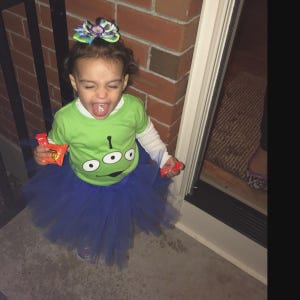 May include: A child in a Halloween costume, wearing a green t-shirt with an alien face graphic, a blue tutu, and a colorful hair bow. The child is holding candy and has their mouth open.