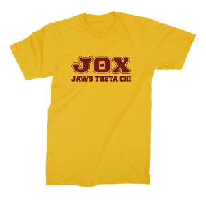 Peut inclure: Un t-shirt jaune avec un motif marron qui dit "JOX JAWS THETA CHI"