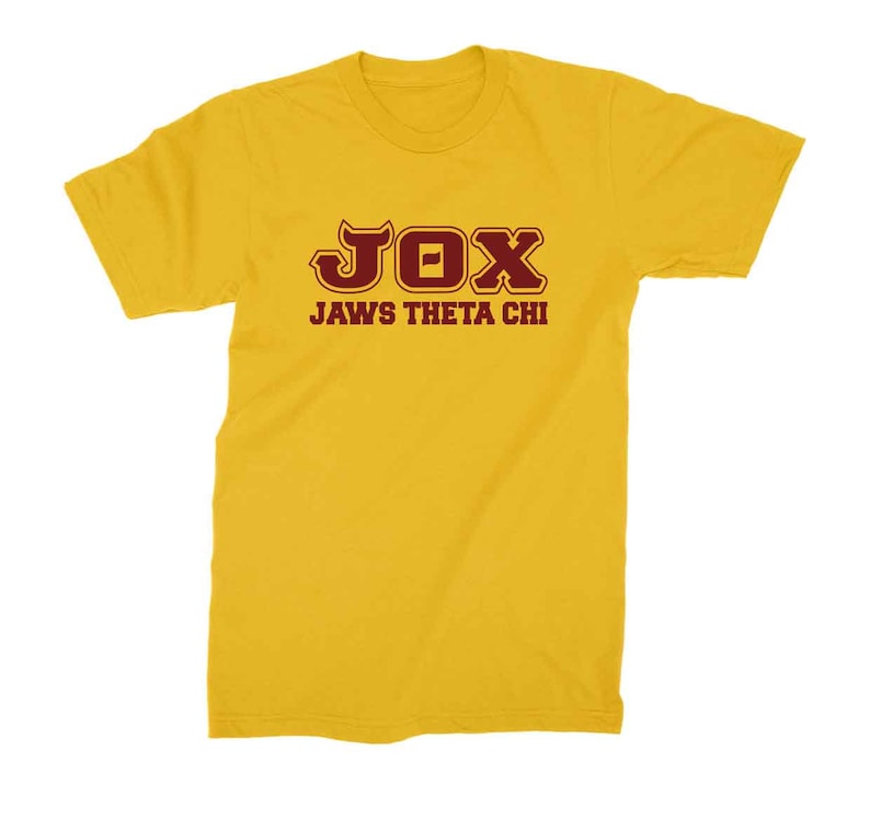 Peut inclure: Un t-shirt jaune avec un motif marron qui dit "JOX JAWS THETA CHI"