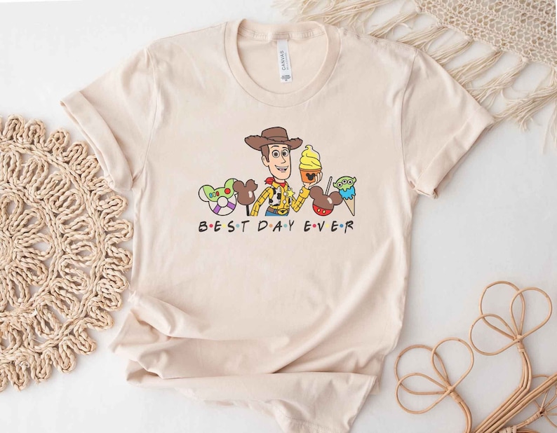 May include: A beige t-shirt with a cartoon image of Woody from Toy Story, Mickey Mouse ears, and a green alien. The text "BEST DAY EVER" is printed below the image.