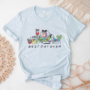 May include: Light blue t-shirt with a graphic of Buzz Lightyear from Toy Story and Mickey Mouse characters. The text "BEST DAY EVER" is printed below the graphic.