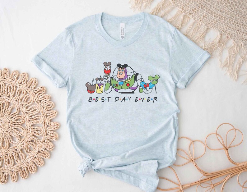 May include: Light blue t-shirt with a graphic of Buzz Lightyear from Toy Story and Mickey Mouse characters. The text "BEST DAY EVER" is printed below the graphic.