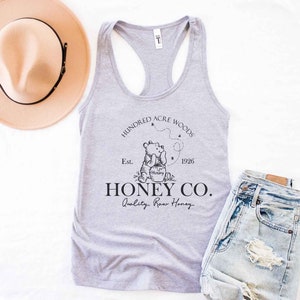 May include: A gray tank top with a black graphic of Winnie the Pooh holding a jar of honey. The graphic also includes the text "Hundred Acre Woods", "Est. 1926", "Honey Co.", and "Quality Raw Honey".