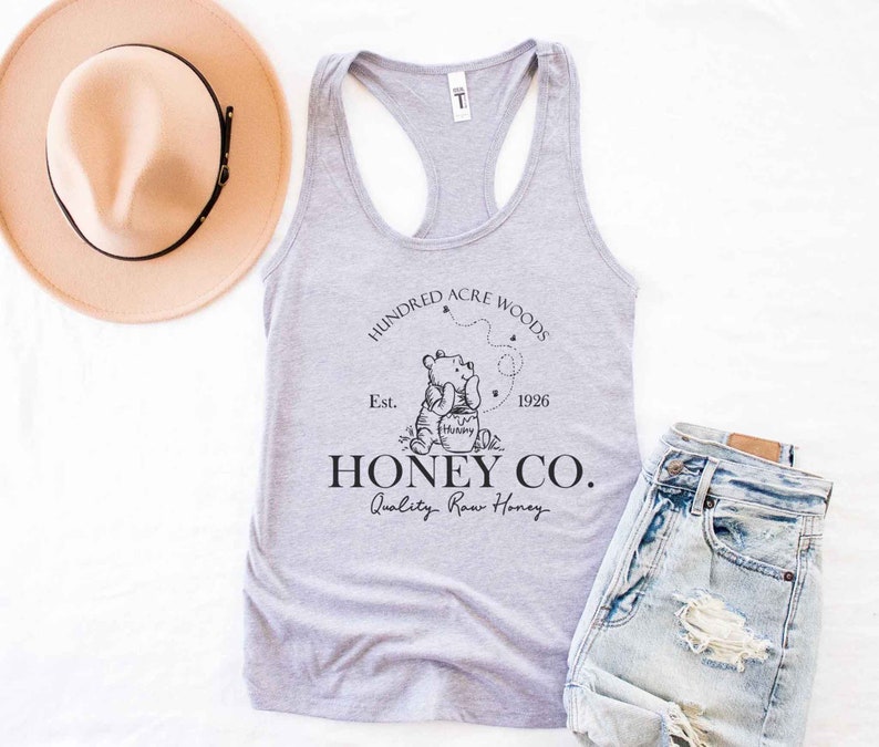 May include: A gray tank top with a black graphic of Winnie the Pooh holding a jar of honey. The graphic also includes the text "Hundred Acre Woods", "Est. 1926", "Honey Co.", and "Quality Raw Honey".