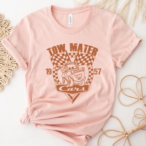 以下が含まれることがあります： 映画「カーズ」のキャラクター、メーターのブラウンのグラフィックがプリントされたピンクのTシャツ。グラフィックには、「Tow Mater」、「Cars」、「19」、「57」の文字が含まれています。