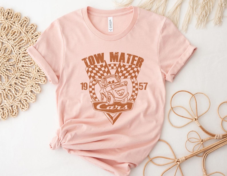 以下が含まれることがあります： 映画「カーズ」のキャラクター、メーターのブラウンのグラフィックがプリントされたピンクのTシャツ。グラフィックには、「Tow Mater」、「Cars」、「19」、「57」の文字が含まれています。
