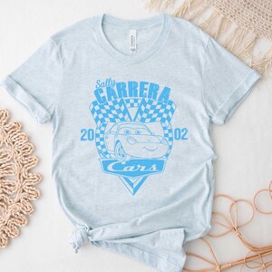 以下が含まれることがあります： 薄い青色のTシャツに、映画「カーズ」のサリー・カーレラをモチーフにした青いグラフィックがプリントされています。グラフィックには、「サリー・カーレラ」、「2002」、「カーズ」の文字が書かれています。