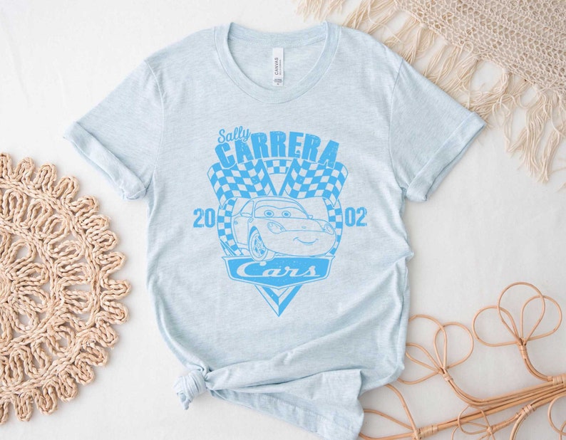 以下が含まれることがあります： 薄い青色のTシャツに、映画「カーズ」のサリー・カーレラをモチーフにした青いグラフィックがプリントされています。グラフィックには、「サリー・カーレラ」、「2002」、「カーズ」の文字が書かれています。