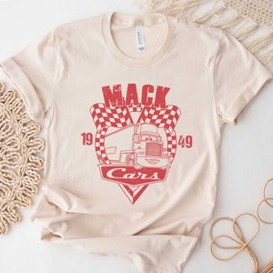 以下が含まれることがあります： ベージュのTシャツに、赤いトラックのグラフィックと「Mack Cars 19 49」の文字がプリントされています。