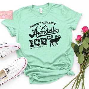 Puede incluir: Una camiseta verde turquesa con un dise&ntilde;o gr&aacute;fico negro que dice "Finest Quality Arendelle Ice Co. Established 2013". El gr&aacute;fico tambi&eacute;n incluye un par de picos cruzados y una silueta de un ciervo.