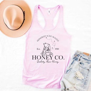 May include: Pink tank top with a black graphic of Winnie the Pooh holding a jar of honey. The graphic also includes the text "Hundred Acre Woods", "Est. 1926", and "Honey Co. Quality Raw Honey".