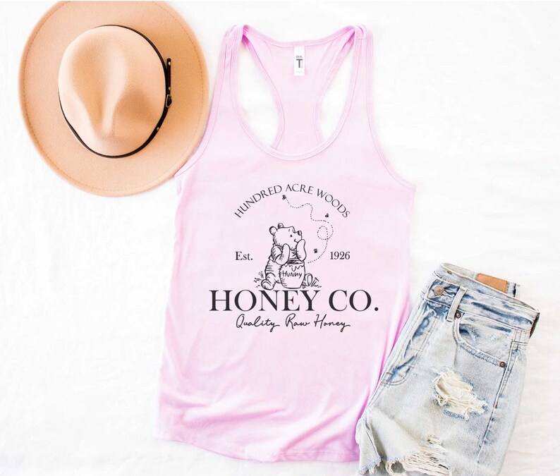 May include: Pink tank top with a black graphic of Winnie the Pooh holding a jar of honey. The graphic also includes the text "Hundred Acre Woods", "Est. 1926", and "Honey Co. Quality Raw Honey".