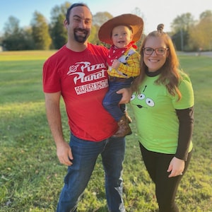 May include: A family dressed in costumes poses outdoors. The father wears a red Pizza Planet t-shirt, the child is dressed as Woody from Toy Story, and the mother wears a green shirt with an alien graphic. The background is a grassy field.